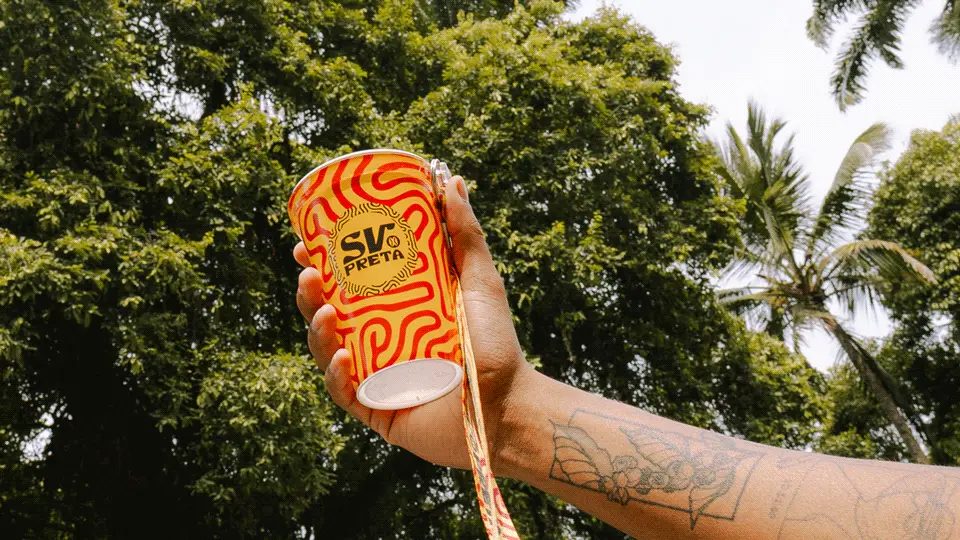 Mão segurando copo laranja e amarelo com a marca SV Preta diante de árvores