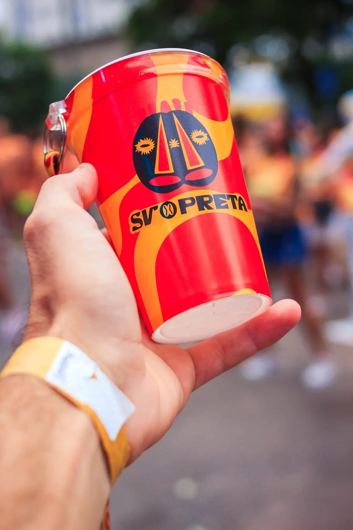 Mão com pulseira segurando copo vermelho com o emblema do SV Preta em meio à festa