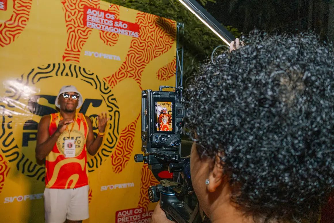 Pessoa de abadá laranja posando diante do backdrop do SV Preta enquanto é filmada