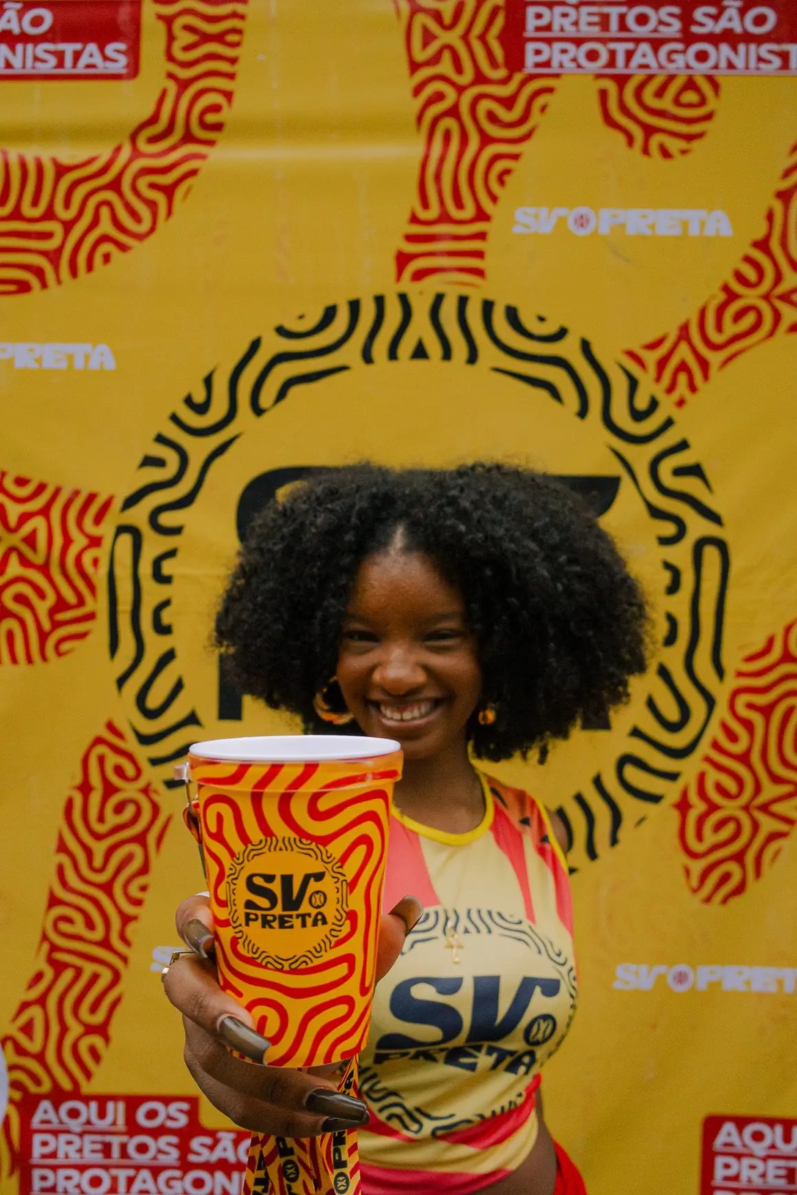 Mulher sorrindo diante de painel laranja do SV Preta, segurando um copo da marca