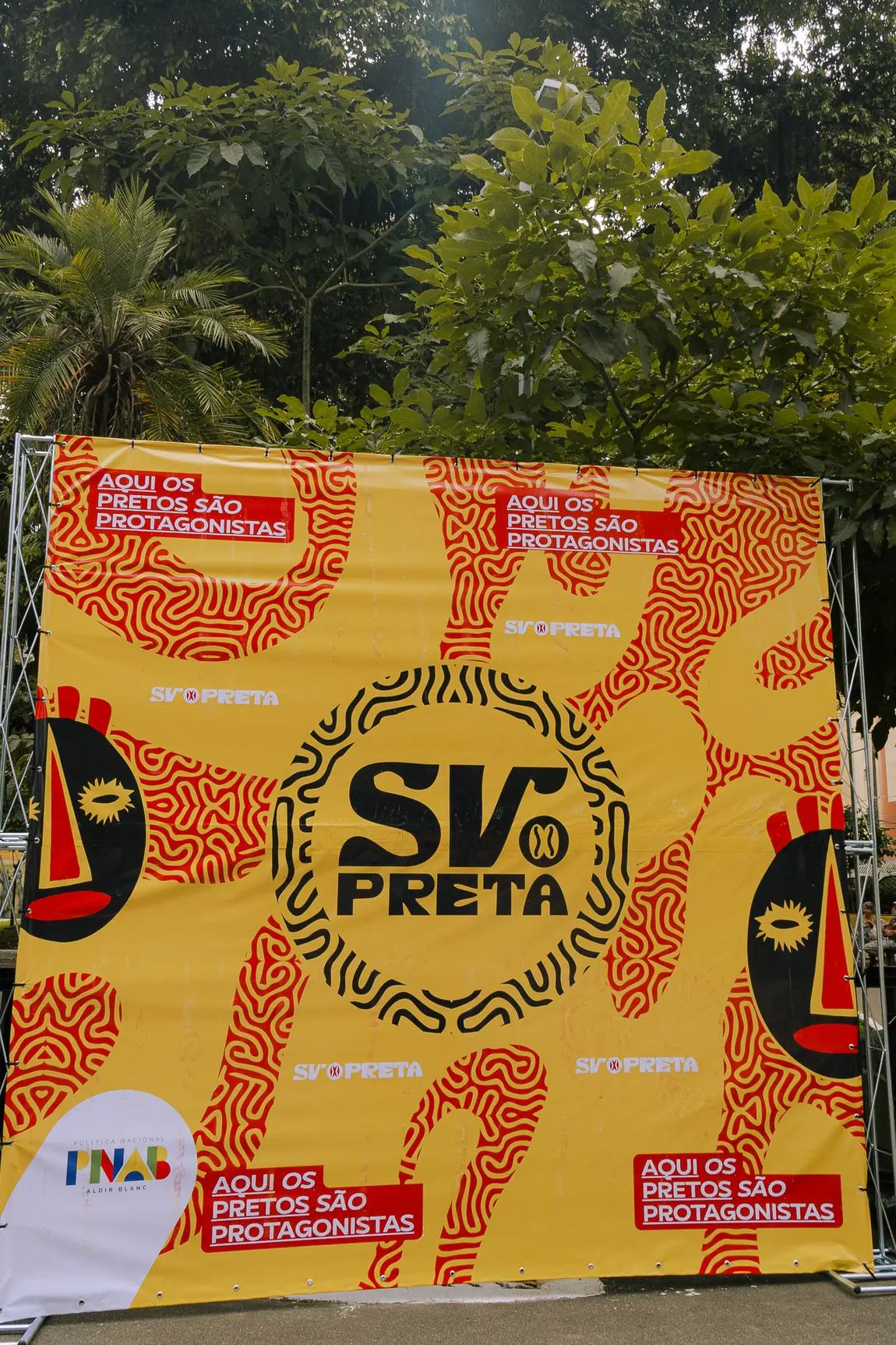 Backdrop laranja e amarelo do SV Preta com o grafismo e o emblema, em ambiente aberto